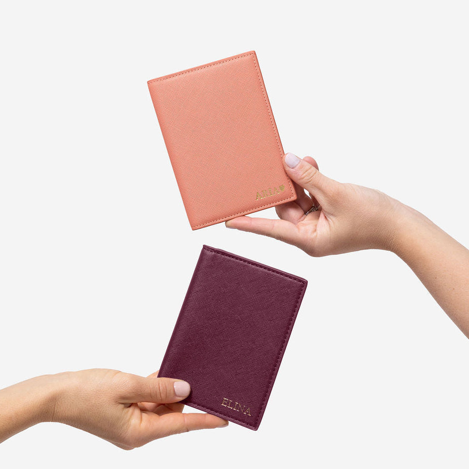 Two hands hold up personalized Saffiano leather passport holders: one peach with 'ARIA' and one burgundy with 'ELINA' initials.