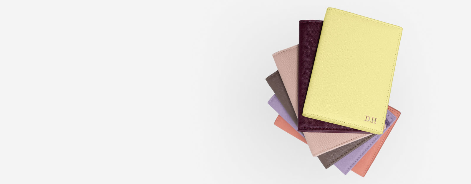 Stack of Saffiano leather passport holders in various colors: yellow, burgundy, pink, khaki, purple, and peach. The top yellow cover has D.H. embossed.