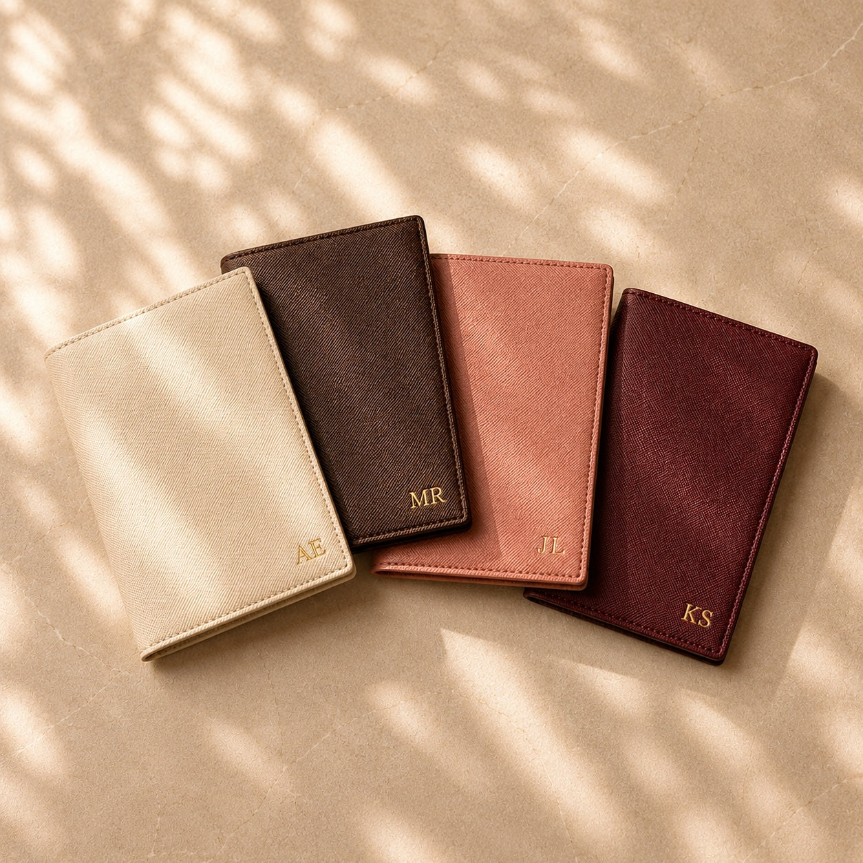 Four leather wallets in different colors with initials on a beige surface with shadow patterns.